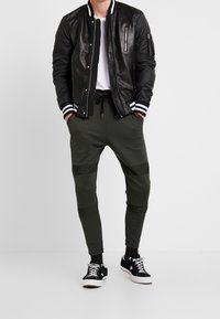 Black leather bomber jacket with striped cuffs, white t-shirt, olive green joggers with panel details, and black sneakers with white soles.