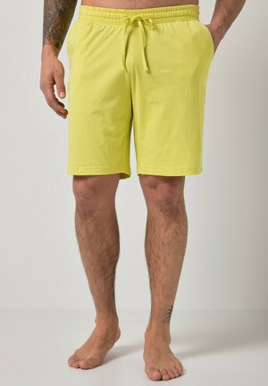 Mężczyzna w jaskrawożółtych szortach ze sznurkiem, stoi boso, z jedną ręką w kieszeni i tatuażem na lewym przedramieniu oraz prawym kostce.