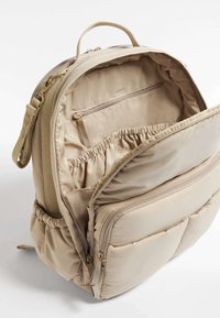Beige ryggsäck gjord av mjukt tyg med flera fack, inklusive ett dragkedjefack och elastiska sektioner. Vadderad design med ett handtag.