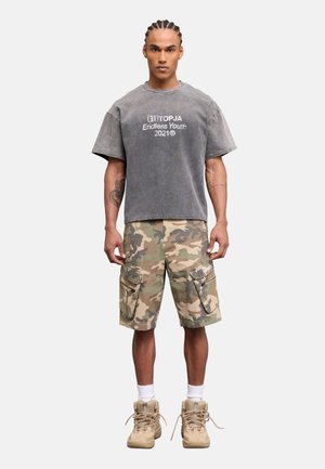 Hombre con trenzas, con camiseta gris desgastada "TOPJA Endless Youth 2021", pantalones cortos cargo de camuflaje, calcetines blancos y botas beige, de pie erguido.