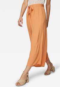 Broek in oranje stof met een relaxed fit, voorzien van een gestrikte taille en plooien. Gecombineerd met beige sandalen met banden, die de enkel accentueren.