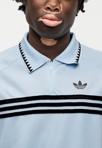 Ανοιχτό μπλε πόλο με φερμουάρ στο γιακά, μαύρες ρίγες, υφή διχτυού και μαύρο λογότυπο στο στήθος. Διαθέτει αντίθετο μοτίβο γιακά.