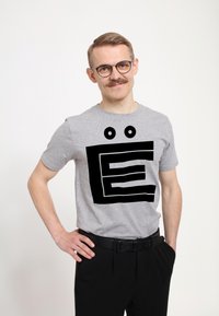 Šedé tričko s krátkým rukávem, které zdobí velká černá grafika připomínající písmeno "E" se dvěma kulatýma očima nad ním. Nosit s černými kalhotami.