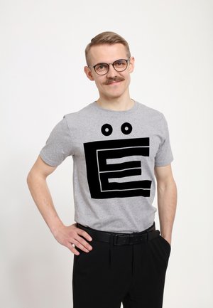 Šedé tričko s krátkým rukávem, které zdobí velká černá grafika připomínající písmeno "E" se dvěma kulatýma očima nad ním. Nosit s černými kalhotami.
