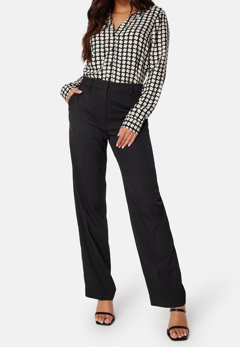 Femme portant un chemisier à manches longues noir et blanc à motifs, un pantalon noir taille haute et des talons ouverts noirs.
