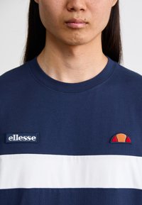 Marineblåt bomulds T-shirt med en hvid vandret stripe, der har et broderet "ellesse" logo og et lille farvet emblem på brystet.