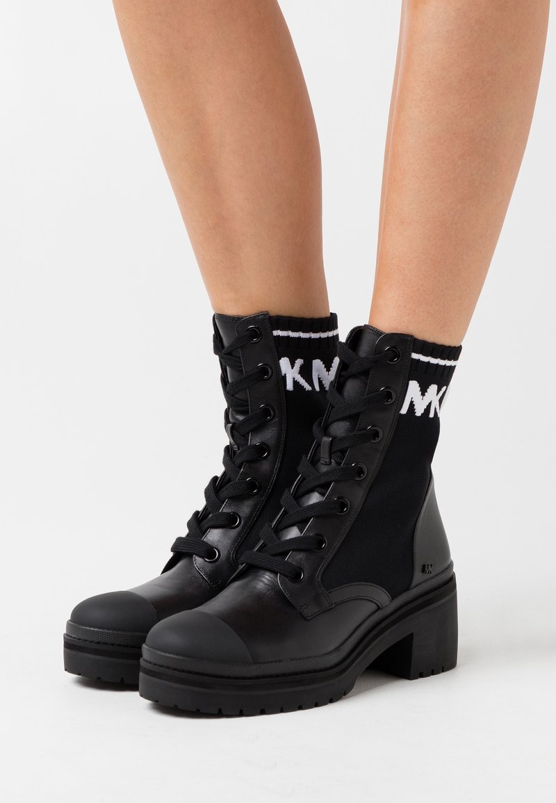 Bottes noires à lacets, à talons épais et chevilles en maille semblables à des chaussettes avec un lettrage blanc, portées par une personne debout sur une surface blanche.