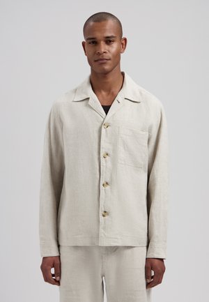 Mand med kort hår iført en beige langærmet skjorte med knapper og brystlomme, stående mod en ensartet lysegrå baggrund.