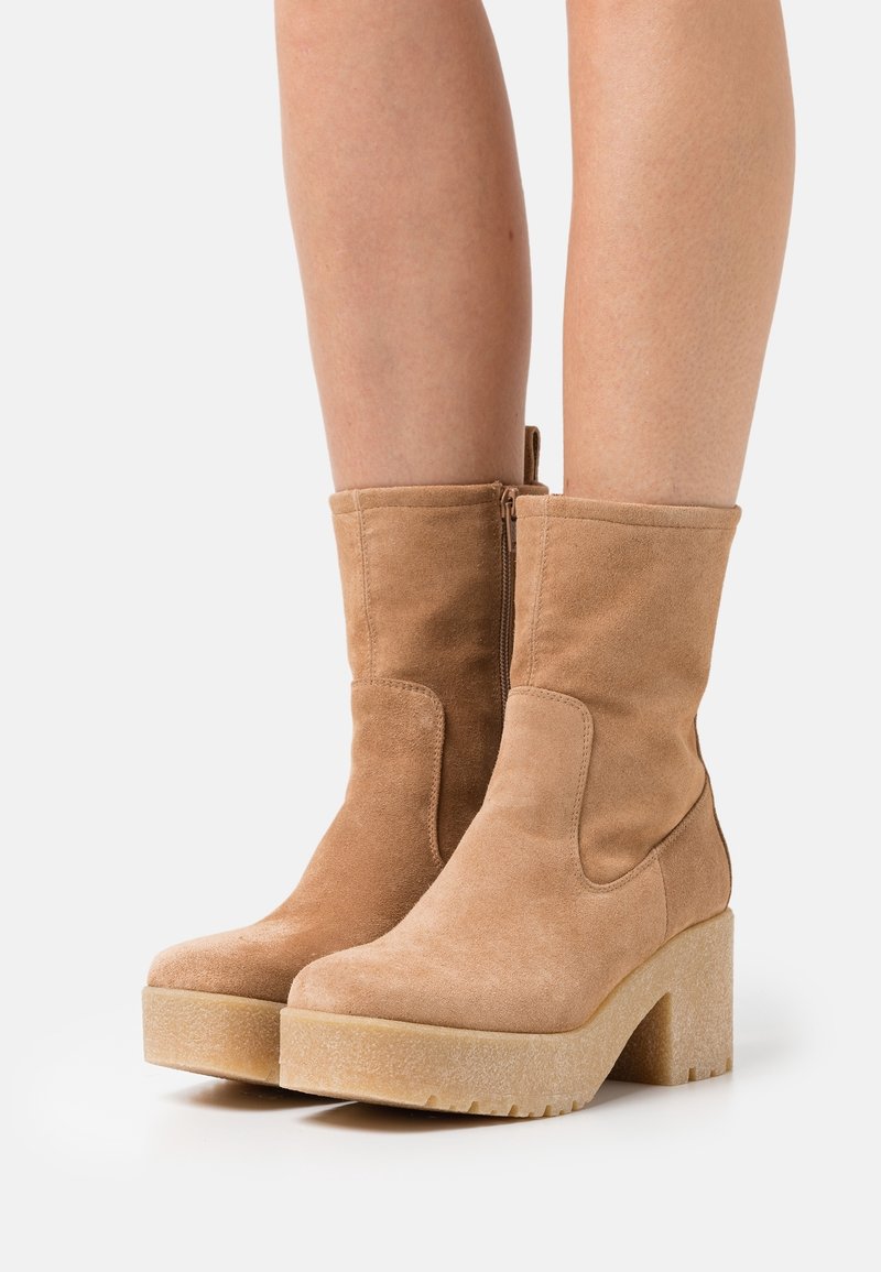 Bottes mi-mollet en suède beige avec talons plateforme épais et fermetures éclair sur les côtés, portées sur jambes nues sur un fond uni.