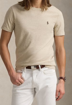 Mand iført beige kortærmet T-shirt med lille logo, hvide bukser, brun bælte og brun ursrem, hånd i lommen.