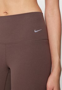 Legíny vyrobené z elastického materiálu v bohatej hnedej farbe s hladkým povrchom. Majú malé šedé logo Nike na páske.