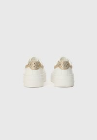 Sneakers bianche con un accento di glitter oro a effetto tessuto nel tallone, dotate di una suola spessa e materiale liscio, progettate per l'uso quotidiano.