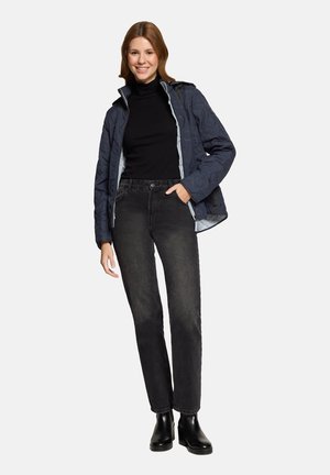 Young woman smiling, wearing a navy quilted jacket, black turtleneck, faded black jeans, and black ankle boots, standing with hand in pocket.