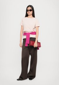 Woman wearing beige t-shirt, wide dark pants, pink sweater tied at waist, sunglasses, holding red clutch bag against plain background.