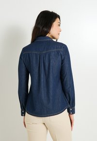 Camisa de mezclilla en azul oscuro con mangas largas, cuello de camisa y puños abotonados. Presenta un yugo trasero y detalles de costura sutiles.