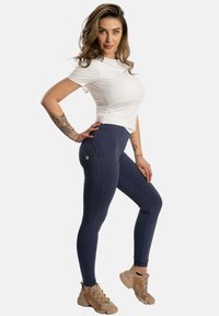 Marineblaue High-Waist-Leggings mit glatter Textur und gestickten Details, kombiniert mit einem taillierten weißen T-Shirt und beigen Sportschuhen.