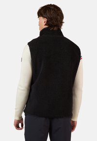 Gilet di pile nero con colletto alto e senza maniche, tessuto testurizzato. Indossato sopra una camicia bianca a maniche lunghe, con polsini a coste.