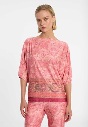 Vrouw die een loszittende top met roze patroon draagt en bijpassende broek met een gedetailleerd horizontaal bandontwerp over de taille.