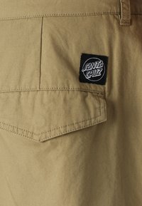 Beige fabric shorts featuring a back pocket with an angled flap and a black label with "SANTA CRUZ" logo in white. Soft texture, casual design.