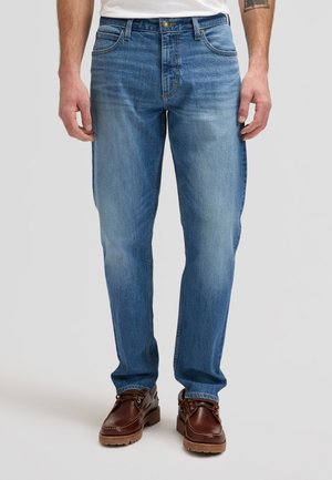 Man draagt lichtblauwe recht gesneden spijkerbroek en bruine leren laarzen, staand tegen een effen lichte achtergrond.