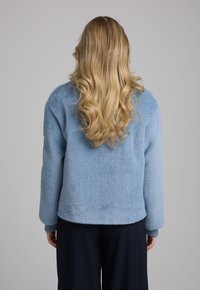 Lichtblauwe fleecetrui met een gestructureerde afwerking, voorzien van een ronde hals en lange mouwen, gedragen met donkere broeken.