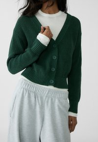 Green knitted cardigan with a v-neck, five-button front, and ribbed cuffs. Worn over a white long-sleeve top and paired with gray sweatpants.