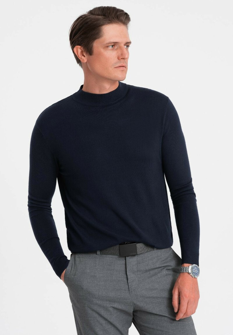 Marineblauer langärmeliger Strickpullover mit hohem Kragen, kombiniert mit grauen Anzughosen und einer silbernen Uhr, vor einem weißen Hintergrund platziert.