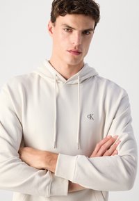 Cremfarbener Hoodie aus Baumwollmischung mit Kordelzugkapuze, gerippten Bündchen und kleinem gesticktem Logo auf der Brust. Glatte Textur.