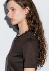 Top marrone aderente con maniche corte, dotato di un colletto rotondo accentuato da un dettaglio in catena argentata. La modella ha capelli lunghi e ondulati.