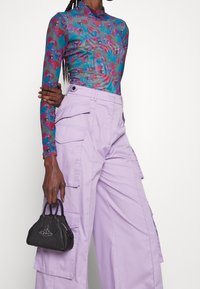 Mehrfarbige, transparente Langarm-Bluse mit floralen Mustern, kombiniert mit hoch taillierten, lilafarbenen Cargo-Hosen und einer kleinen schwarzen Handtasche.