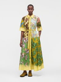 Mannequin portant une longue robe patchwork florale ceinturée, à manches courtes et col, associée à des chaussures jaune moutarde.