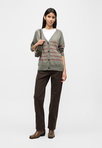 Γυναίκα που φοράει γκρι ζακέτα με κόκκινες ρίγες, λευκό πουκάμισο, σκούρο καφέ παντελόνι, καφέ μποτάκια και κρατάει χακί πράσινη τσάντα ώμου.