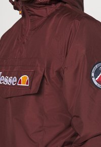 Tummanpunainen vetoketjullinen pusero, jossa etutasku ja kirjailtuja logokoristeita. Sileä, kevyt materiaali.