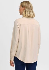 Blusa beige a maniche lunghe in tessuto morbido e leggero. Presenta un orlo arrotondato, dettagli plissettati sul retro e polsini con bottoni.
