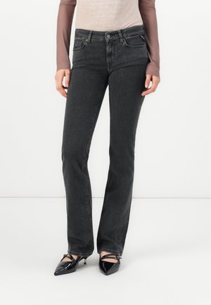 Frau trägt hoch taillierte, dunkelgraue Jeans mit geradem Bein und schwarze spitze Schuhe mit Knöchelriemen, steht vor weißem Hintergrund.