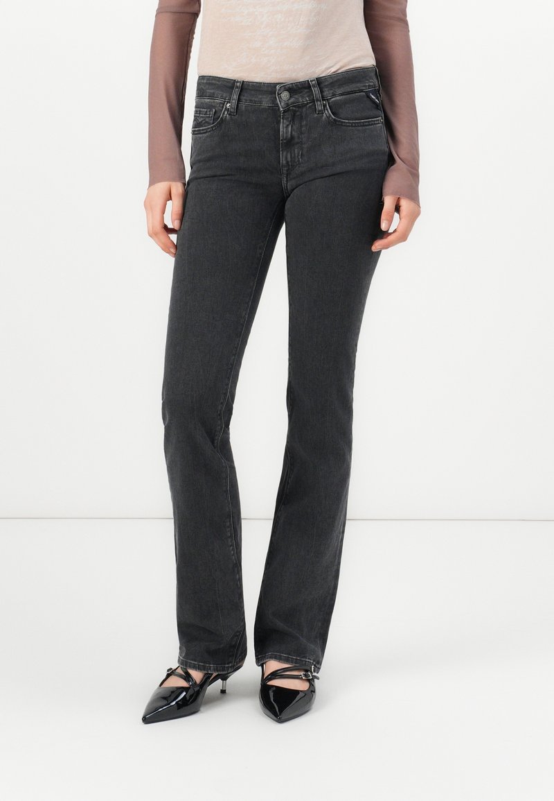 Femme portant un jean gris foncé taille haute à jambes droites et des escarpins noirs à bout pointu avec des brides à la cheville, debout devant un fond blanc.