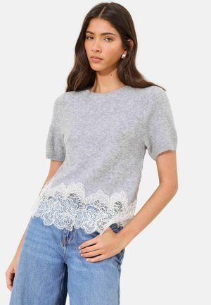 Young woman with long dark hair wearing a short-sleeve grey sweater with white lace trim and light blue jeans, posing with one hand in pocket.