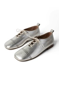 Par de zapatos de cuero plateado metálico con cordones, diseño perforado en la puntera y tacones bajos marrones, exhibidos sobre fondo blanco.