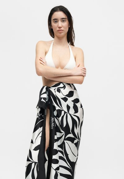 Γυναίκα όρθια με σταυρωμένα χέρια, φοράει λευκό τοπ μπικίνι με δέσιμο στο λαιμό και μία ασπρόμαυρη παρεό με σχέδιο φύλλων δεμένη στη μέση.