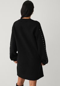 Sort oversized sweaterkjole med tekstureret stof, drop skuldrer og dekorative knapper langs ærmerne. Kantlinjen slutter over knæet.