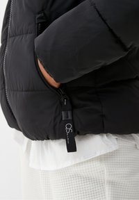 Czarna kurtka puchowa o pikowanej strukturze z boczną kieszenią na zamek i logo na suwaku. Noszona na białej koszuli i wzorzystych spodniach.