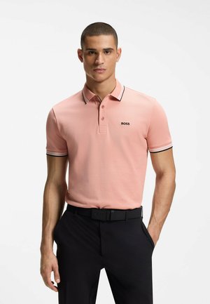 Man draagt een roze polo met korte mouwen en het logo "BOSS", zwarte riem en zwarte broek, staand tegen een witte achtergrond.