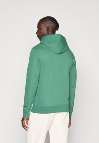 Πράσινο φούτερ με κουκούλα από μαλακό ύφασμα. Διαθέτει θήκη καγκουρό και ριμπ μανσέτες. Απεικονίζεται από πίσω με λείο υφή.