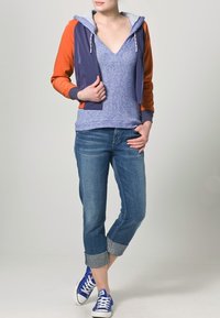 Hoodiejacka med orange och marinblå paneler, över en blå V-ringad tröja, kombinerad med uppvikta blå jeans och blå sneakers.