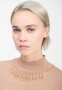Mujer rubia con piel clara que lleva un suéter de cuello alto beige brillante adornado con cuentas de ámbar en forma de lágrima alrededor del cuello.