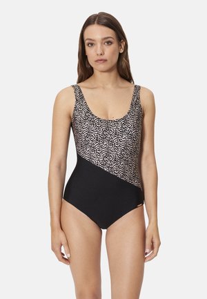 Mujer con un traje de baño de una pieza, con la mitad superior de patrón geométrico y la mitad inferior en negro sólido, de pie contra un fondo blanco.