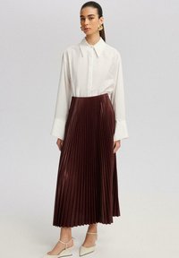 Camicia bianca con maniche ampie abbinata a una gonna plissettata bordeaux, caratterizzata da una texture lucida. Il modello indossa scarpe con tacco bianche.