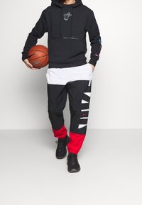 Mann in schwarzem Hoodie und schwarzen, weißen und roten Nike-Hosen, der mit einer Hand einen Basketball hält und nach vorne geht.