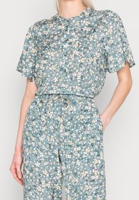 Jumpsuit floreale azzurro chiaro con maniche corte, scollatura rotonda e chiusura anteriore con bottoni. Caratterizzato da motivi di fiori bianchi e gialli e una vita elastica.