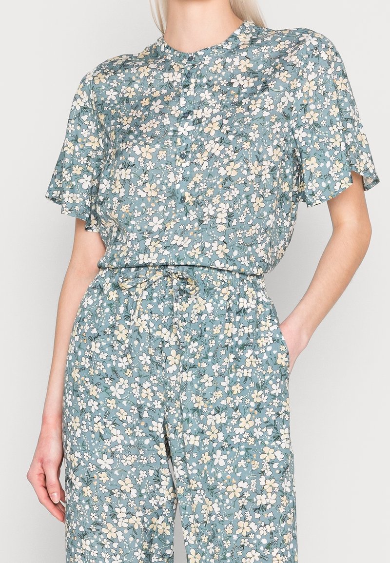 Jumpsuit floreale azzurro chiaro con maniche corte, scollatura rotonda e chiusura anteriore con bottoni. Caratterizzato da motivi di fiori bianchi e gialli e una vita elastica.